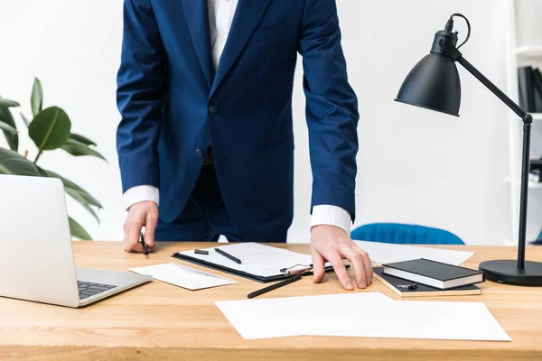 Vue partielle de l'homme d'affaires en costume sur le lieu de travail avec des cahiers, des documents et un ordinateur portable au bureau — Photo
