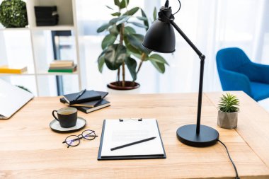 close up view of cup of coffee, eyeglasses, documents and lamp at workplace in office clipart