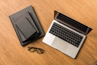 top view of arranged pile of black notebooks, laptop and eyeglasses on wooden tabletop clipart