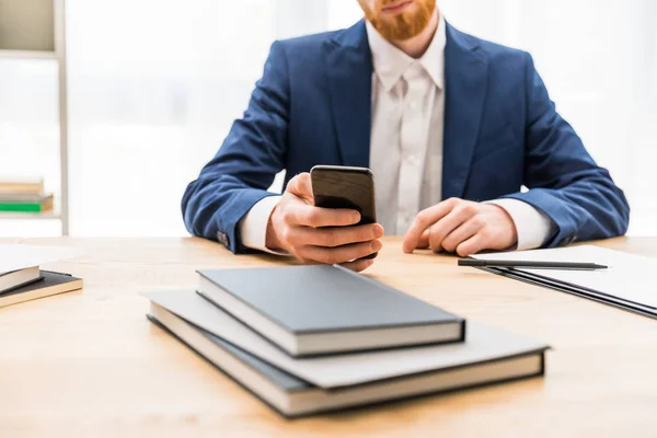 Partial view of businessman in suit using smartphone at workplace with notebooks in office — Stock Photo, Image