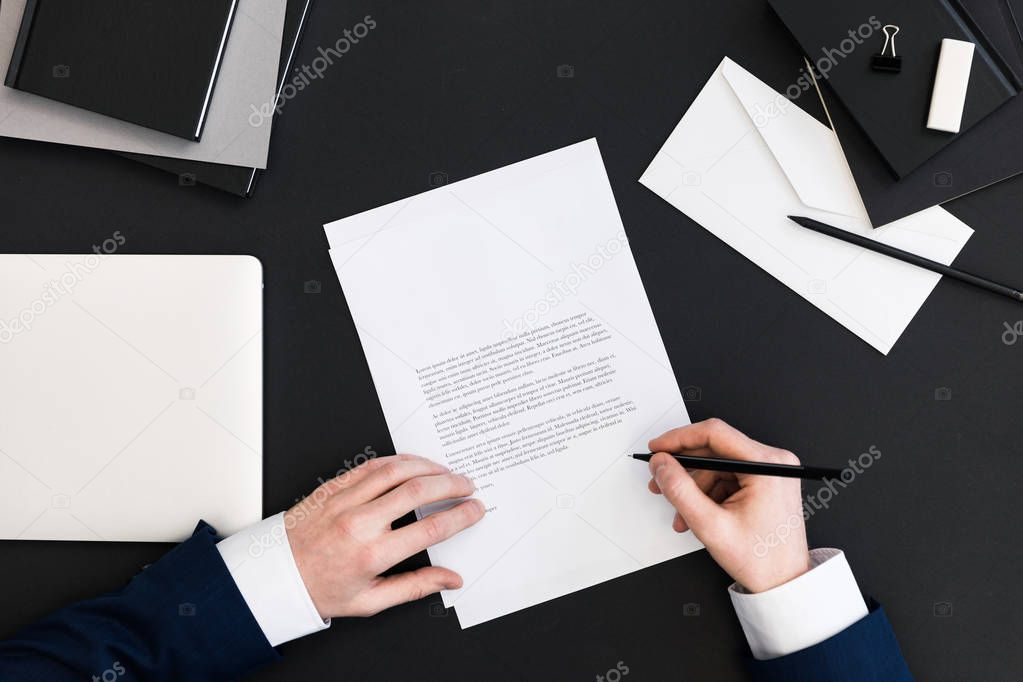 partial view of businessman in suit signing papers at workplace with laptop