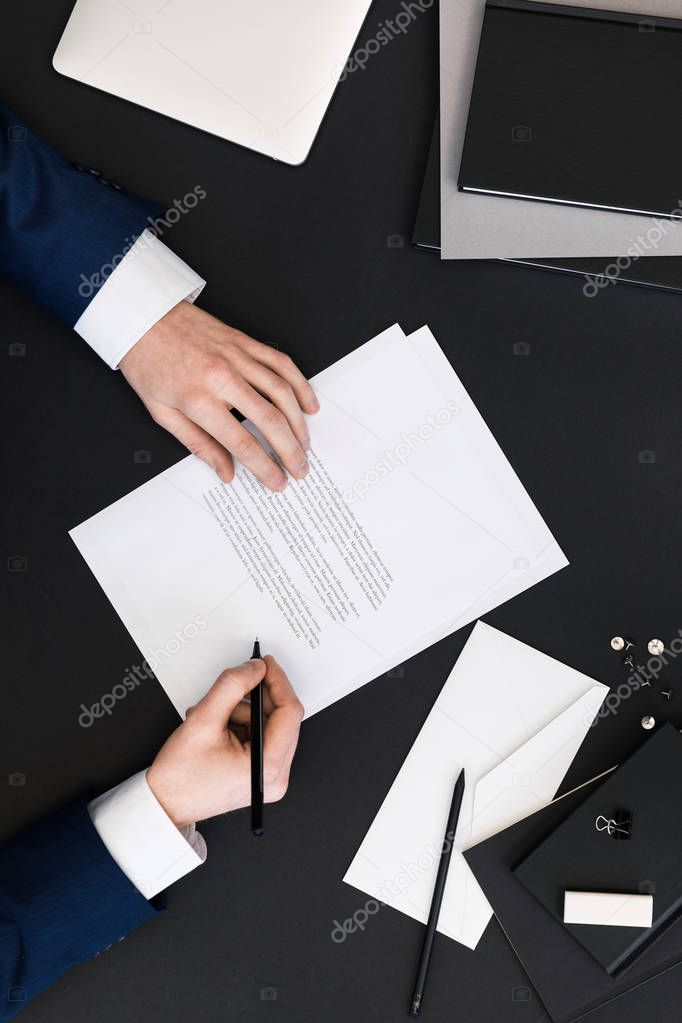 partial view of businessman in suit signing papers at workplace with laptop
