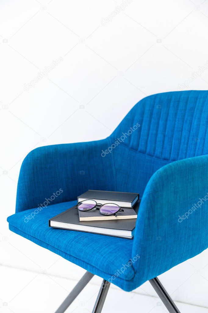 close up view of pile of black notebooks and eyeglasses on blue chair on white backdrop