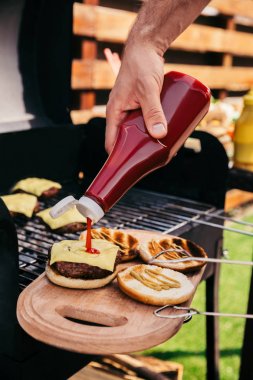 Man adding ketchup to burgers cooked outdoors on grill clipart
