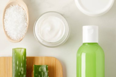 top view of organic cream and shower gel, wooden spoon with salt and aloe vera leaves on cutting board on marble table  clipart