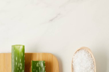 elevated view of aloe vera leaves on cutting board and wooden spoon with salt on marble table clipart