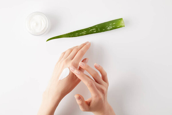 cropped shot of woman applying organic cream on hands, aloe vera leaf and cream container on white surface