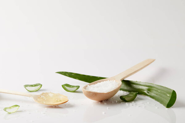 closeup view of two wooden spoons with aloe vera juice and salt, aloe vera leaf and slices on white surface