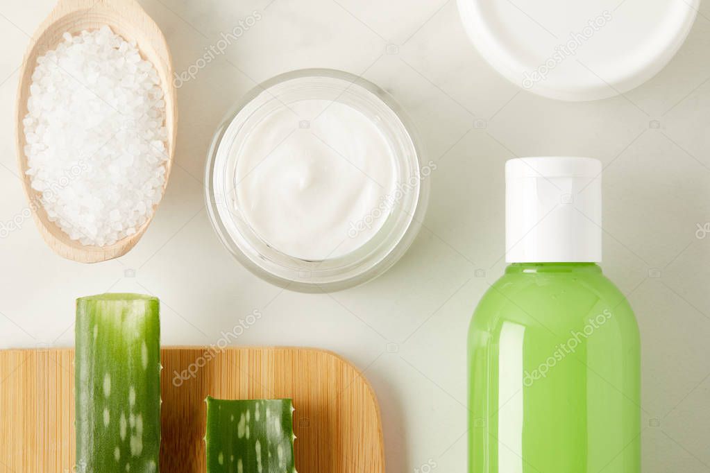 top view of organic cream and shower gel, wooden spoon with salt and aloe vera leaves on cutting board on marble table 