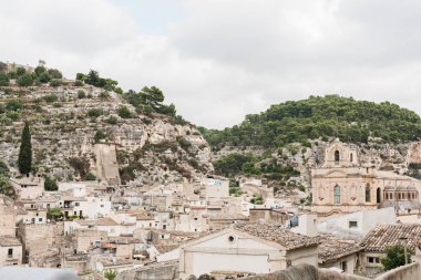 Scicli, İtalya - 3 Ekim 2019: Küçük evleri olan eski bir İtalyan şehri ve bulutlu mavi gökyüzüne karşı kilise 