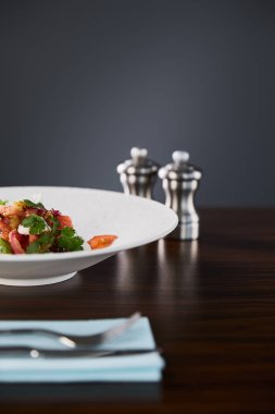 Tahta masadaki beyaz tabakta servis edilen peynirli lezzetli restoran salatasının seçici odak noktası siyah arka planda çatal bıçak, tuz ve biberlik.