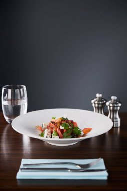Lezzetli restoran salatasının seçici odağı beyaz tabakta servis edilen peynirle ahşap masada su, çatal bıçak takımı ve siyah arka planda tuz ve biber karıştırıcı ile servis ediliyor.
