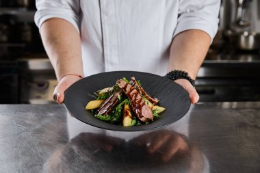 partial view of chef holding dish with arugula, meat and potato at kitchen in restaurant clipart