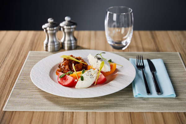 selective focus of delicious restaurant dish with eggplant caviar and tomatoes served on wooden table with water, salt and pepper shakers and cutlery isolated on grey