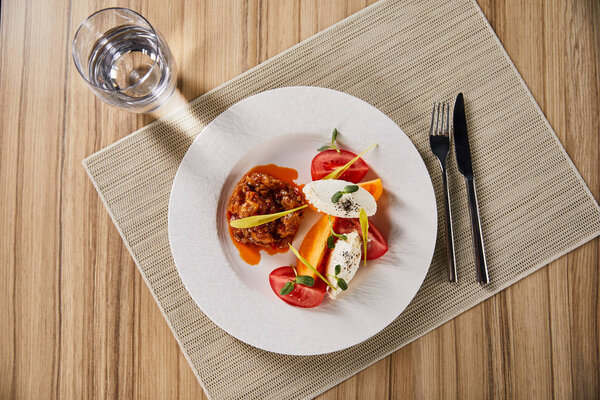 top view of delicious restaurant dish with eggplant caviar and tomatoes served on wooden table with water and cutlery