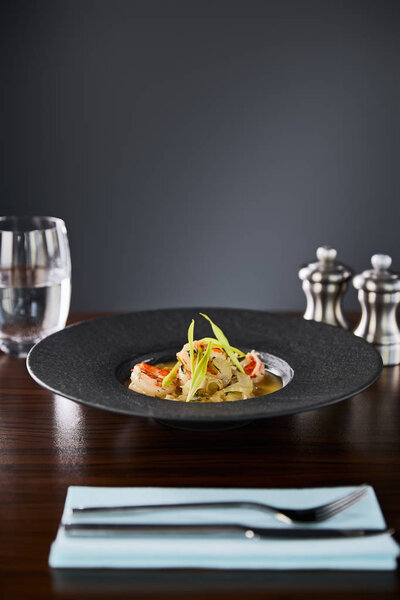 selective focus of delicious restaurant soup with shrimps in black plate served on napkin with cutlery and water on black background