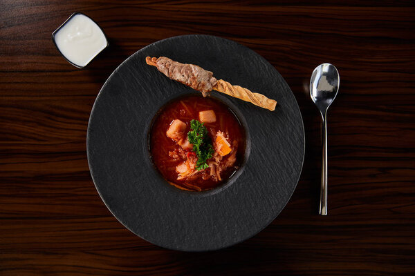 сверху вид на традиционный украинский борщ украшенный петрушкой в черной тарелке подается со сметаной возле ложки за деревянным столом
