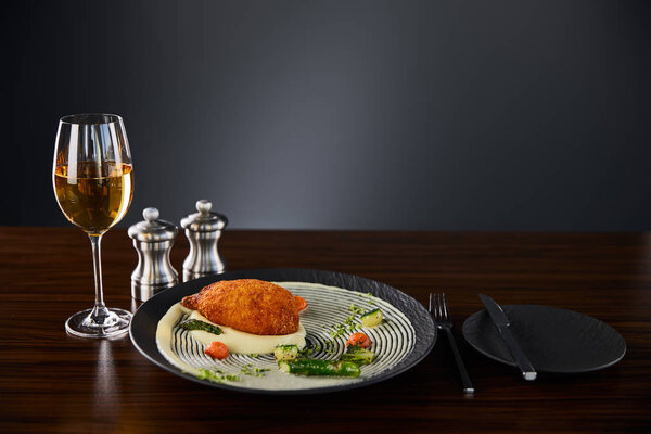 вкусный куриный киев и картофельное пюре подается на тарелке возле столовых приборов и белого вина на черном фоне
