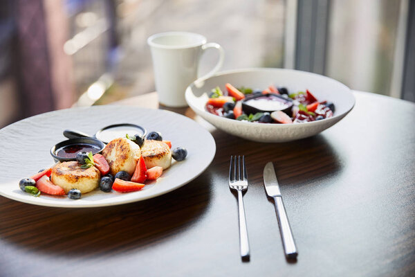 traditional Ukrainian varenyky and syrniki with berries served in white plate with sauce near cutlery and cup on wooden table in restaurant