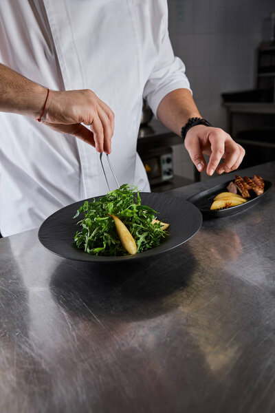 partial view of chef cooking dish with arugula, meat and potato at kitchen in restaurant