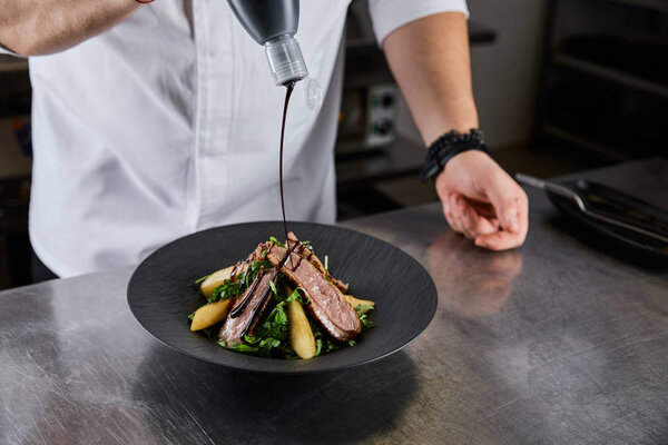 partial view of chef pouring sauce on dish with arugula, meat and potato at kitchen in restaurant