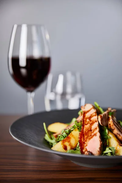 アルグラとおいしい暖かいサラダの選択的な焦点 赤ワインの近くの木製のテーブルの上に黒いプレートのジャガイモと肉 灰色の背景の水 — ストック写真