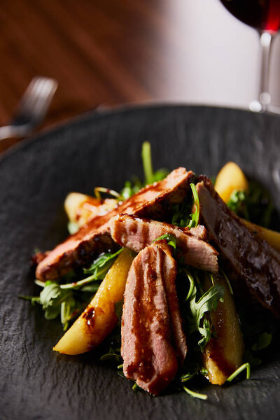 close up view of delicious warm salad with arugula, potato and meat in black plate on wooden table
