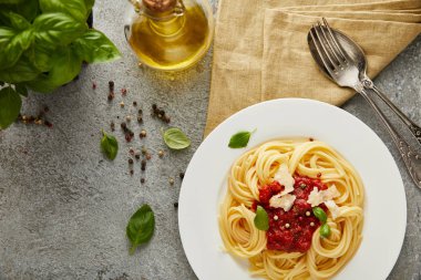 Fesleğen yapraklarının yanında domates soslu lezzetli spagetti, gri desenli yüzeyde yağ ve çatal bıçak seti.