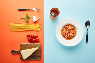 Çatal bıçak takımının yanında domates soslu leziz spagetti ve mavi, kırmızı ve sarı arka planda malzemeler.