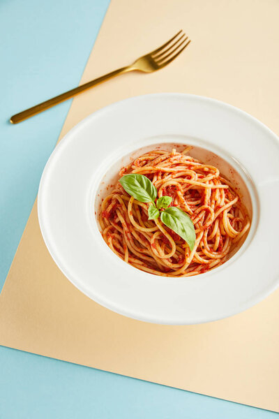 delicious spaghetti with tomato sauce and basil in plate near fork on blue and yellow background