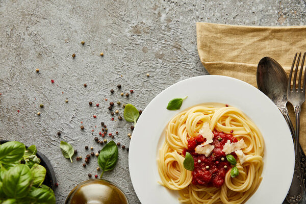 top view of delicious spaghetti with tomato sauce on plate near basil leaves and cutlery on grey textured surface