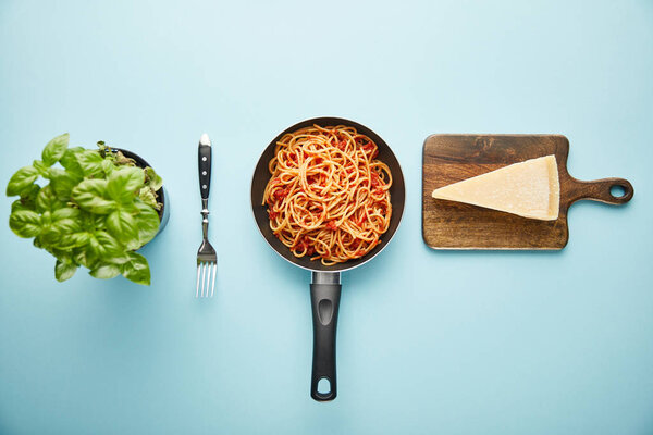 flat lay with delicious spaghetti with tomato sauce in frying pan near basil leaves and parmesan cheese on blue background