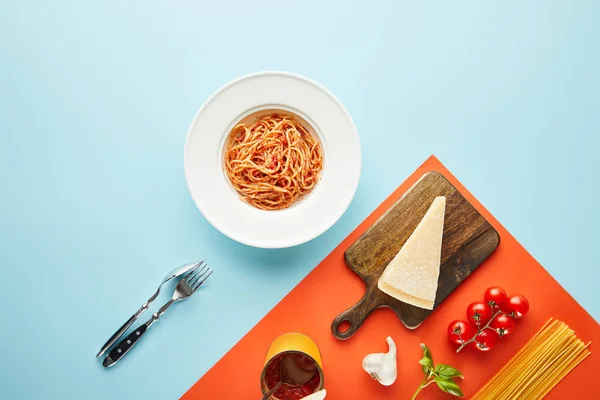 flat lay with delicious spaghetti with tomato sauce in plate near cutlery and ingredients on blue, red background