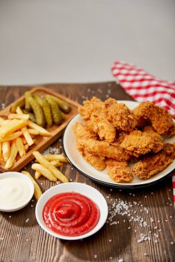 Lezzetli tavuk nugget, ketçap, mayonez, patates kızartması ve salatalık turşusu ahşap masada tuz ve kırsal ekose peçeteyle gri üzerine izole edilmiş.