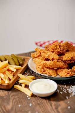 selective focus of delicious chicken nuggets, mayonnaise, french fries and gherkins on wooden table with salt and rustic plaid napkin isolated on grey clipart