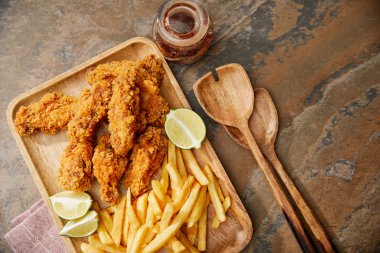 top view of wooden board with delicious chicken nuggets, french fries and lime on stone surface with spatulas clipart