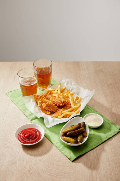 glasses of beer, chicken nuggets with french fries, sauces and gherkins on wooden table on grey background