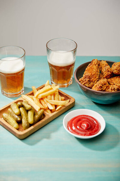 delicious chicken nuggets, ketchup, french fries and gherkins near glasses of beer on turquoise wooden table isolated on grey