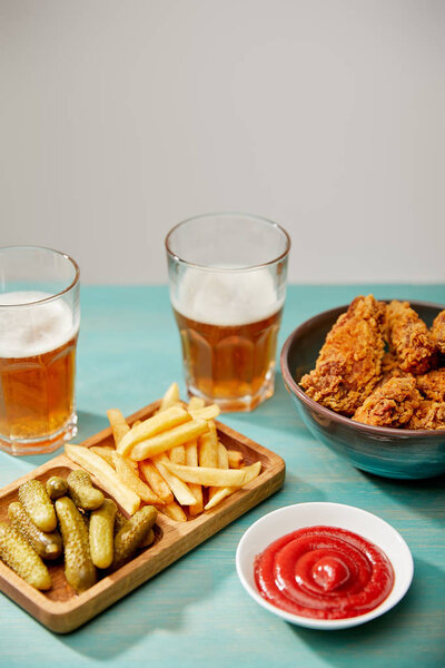 delicious chicken nuggets, ketchup, french fries and gherkins near glasses of beer on turquoise wooden table isolated on grey