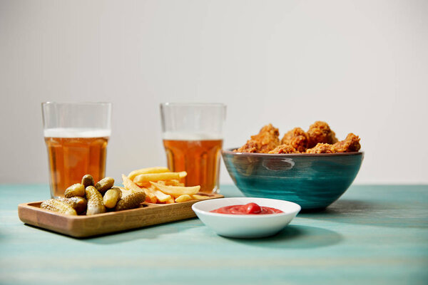delicious chicken nuggets, ketchup, french fries and gherkins near glasses of beer on turquoise wooden table isolated on grey