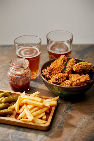 delicious chicken nuggets, french fries and gherkins near glasses of beer on stone surface isolated on grey