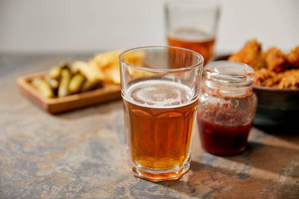 selective focus of delicious chicken nuggets, sauce, french fries and gherkins near glasses of beer on stone surface isolated on grey