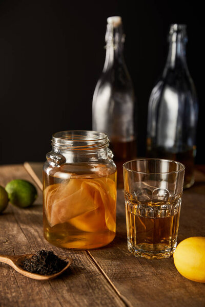 selective focus of glass jar with kombucha near lime, lemon, spice and bottles on wooden table isolated on black