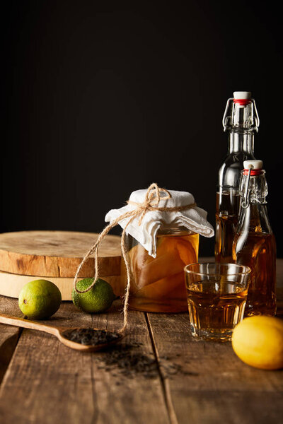glass jar with kombucha near lime, lemon, spice and bottles on wooden table isolated on black