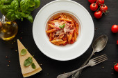 vrchní pohled na chutné boloňské těstoviny s rajčatovou omáčkou a parmezánem v bílém talíři v blízkosti ingrediencí a příborů na černém dřevěném pozadí