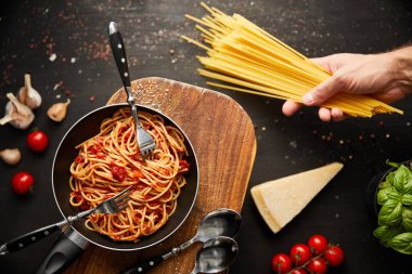 Makarnanın yanında taze malzemelerle birlikte siyah arka planda kızartılmış tavada spagetti tutan bir adam görüntüsü.