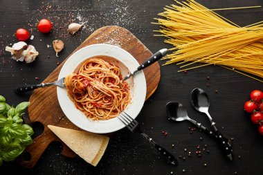 top view of tasty bolognese pasta in white plate near ingredients and cutlery on black wooden background clipart