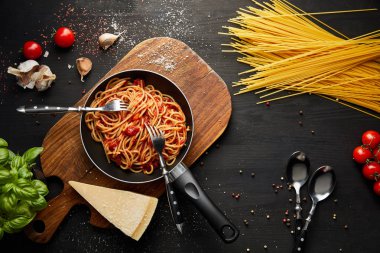 top view of tasty bolognese pasta in frying pan near ingredients and cutlery on black wooden background clipart