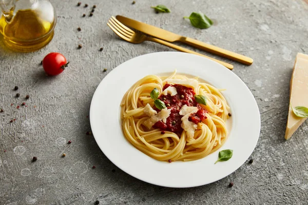 トマトソースのおいしいボロネーゼパスタと材料の近くの白いプレートのパルメザンチーズと灰色の背景のカトラリー — ストック写真