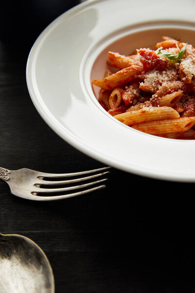 tasty bolognese pasta with tomato sauce and Parmesan in white plate near cutlery on black wooden background